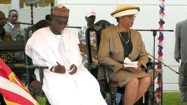 A Black man in a white flowing robe & kufi hat sits next to a Black woman in brown suit & straw hat