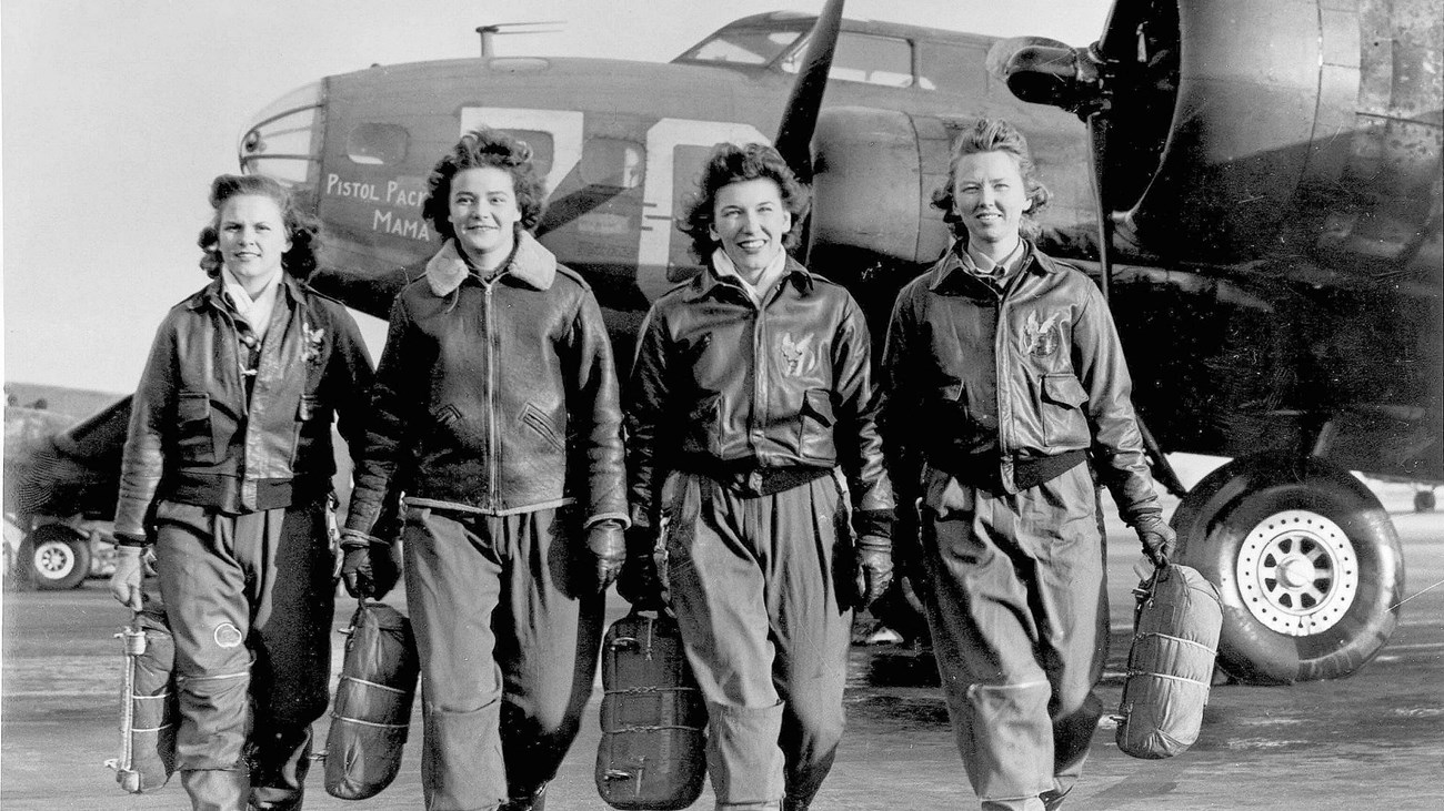 Four women in pilots' attire walk toward the camera with airplane in the background