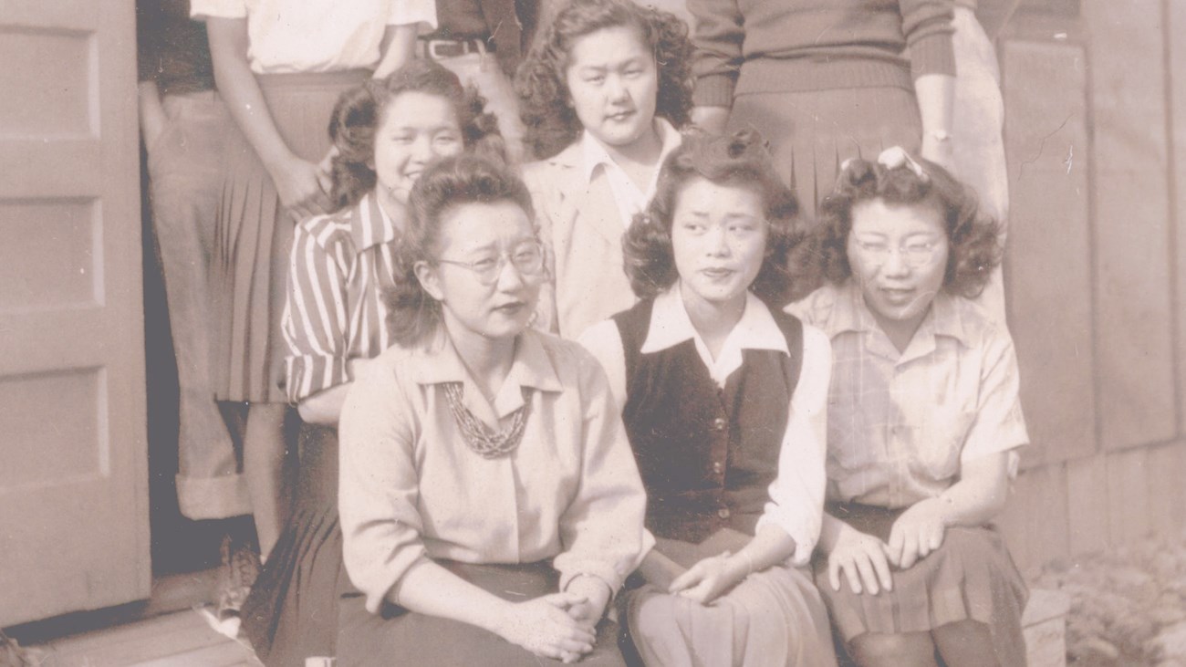 Three young Japanese American women sit on the stoop of a building, looking quizzically at camera