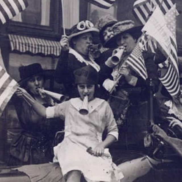Women celebrating by waving American flags and blowing horns. 