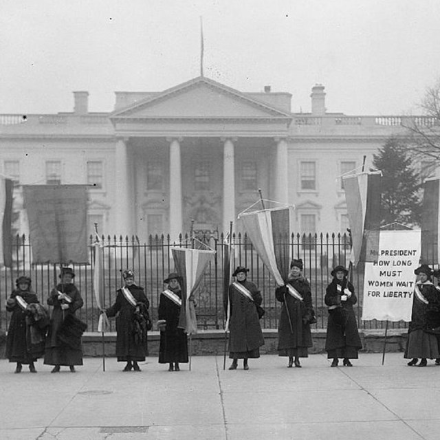 Black and white photo of suffragists holding flags and banners outside White House gates. LOC photo.