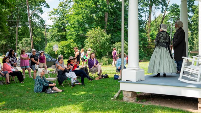 A man and women in 18th century clothing standing on a porch in front of a crowd