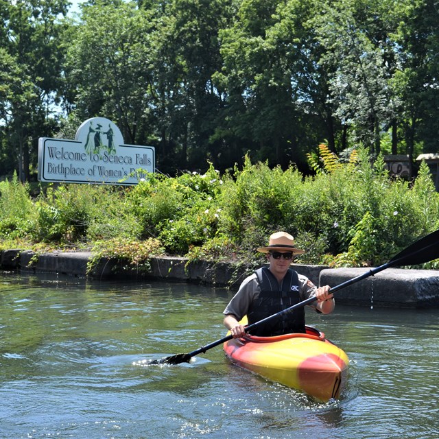 A ranger paddling a kayak past a sign for Seneca Falls