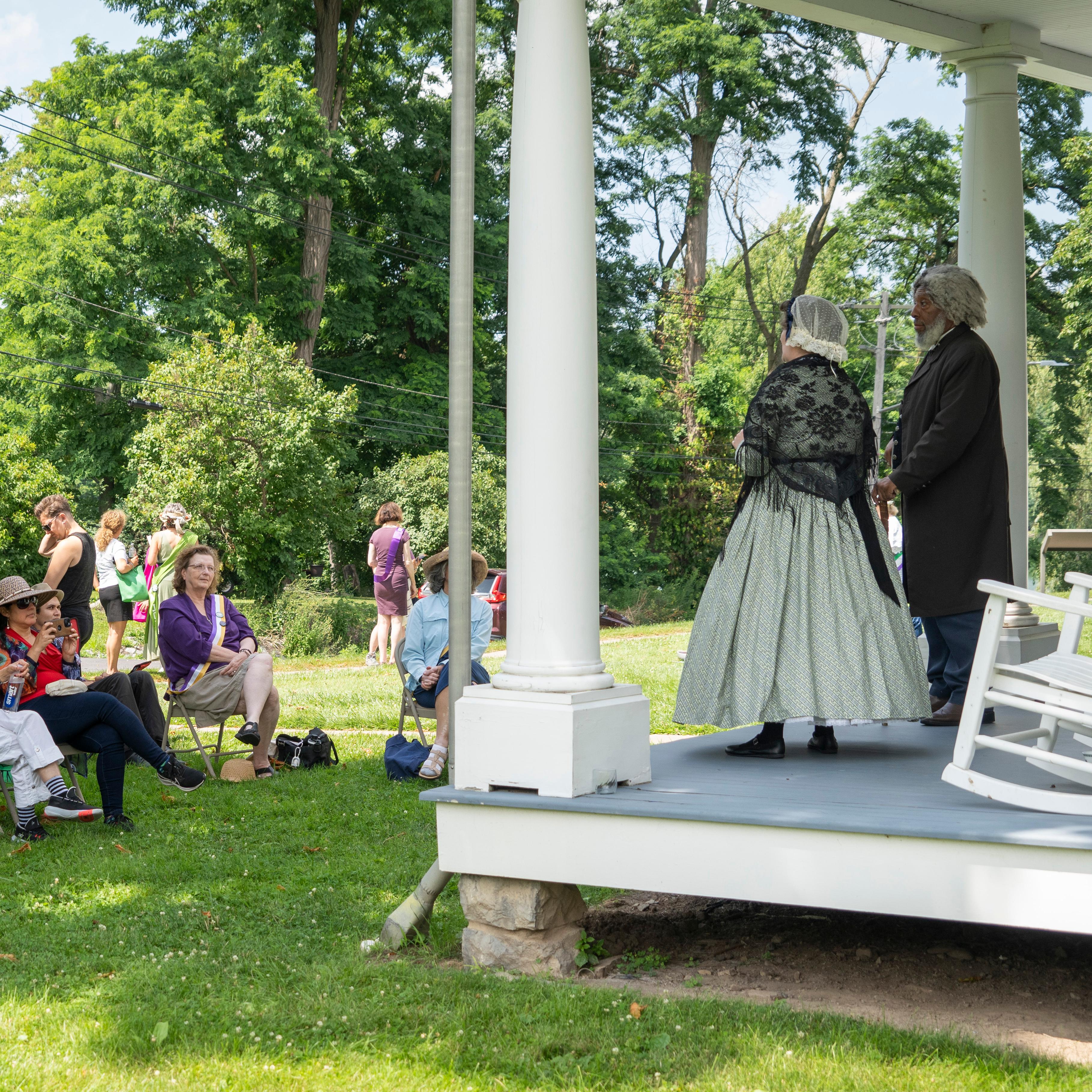 A man and women in 18th century clothing standing on a porch in front of a crowd