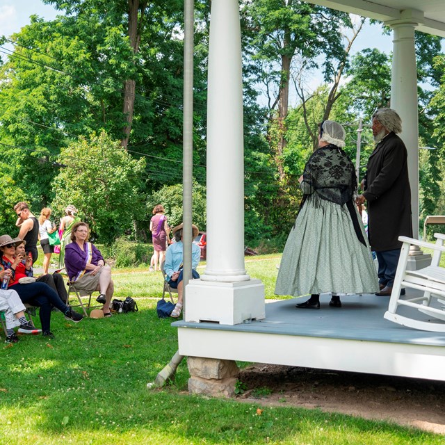 A man and women in 18th century clothing standing on a porch in front of a crowd