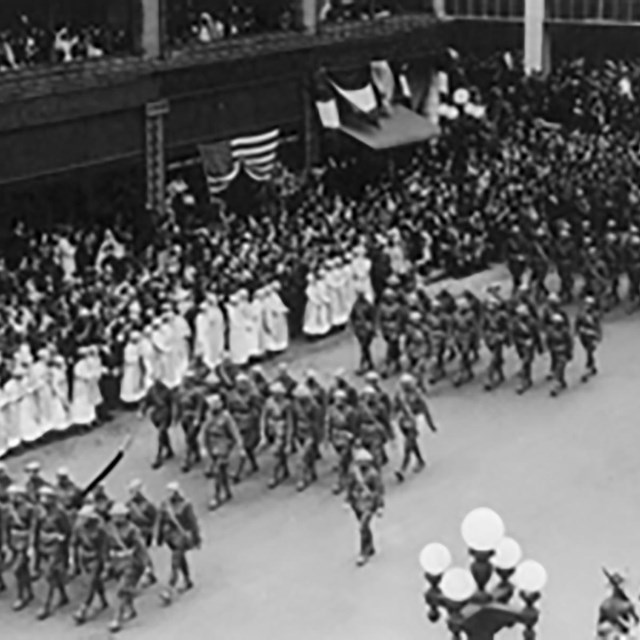 Soldiers march in a parade on a city street lined with spectators.