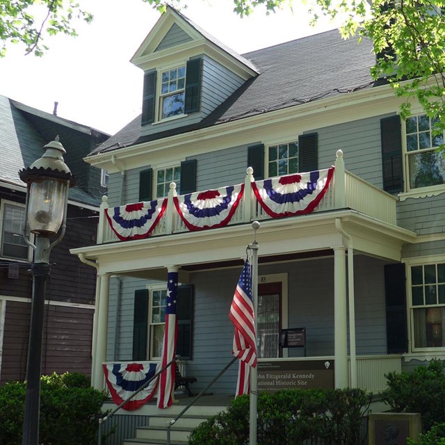 a light blue new england style home with red white and blue bunting on second story porch