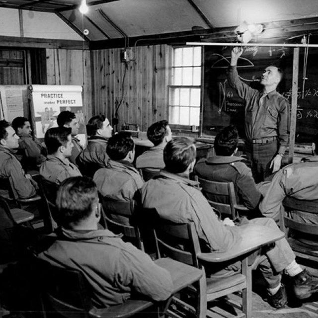 Black and white photo of men sitting in chairs facing instructor and blackboard.