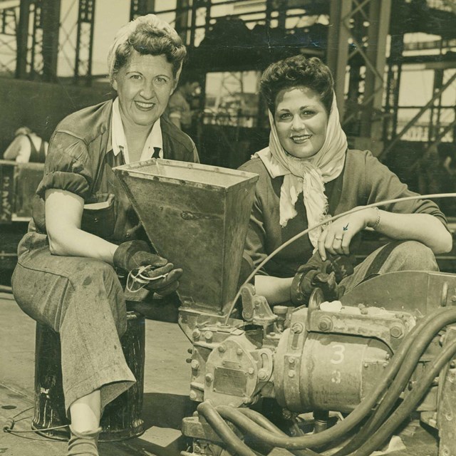 Two women in dungarees site in front of a machine