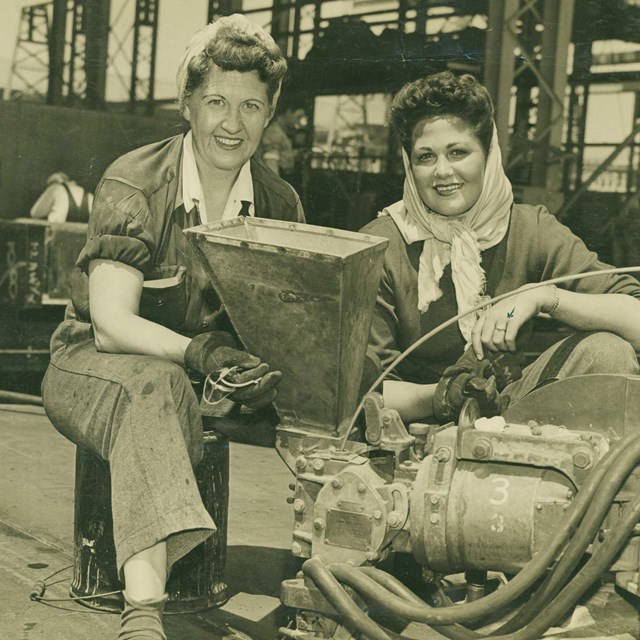 B&W photo of two women in overalls posing in a warehouse