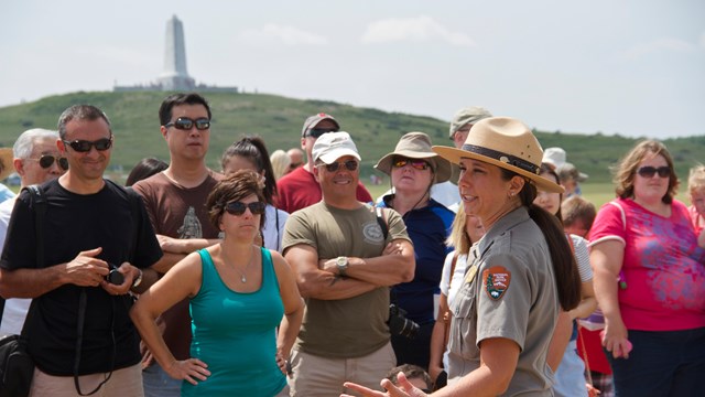 Ranger talks to group of visitors. Green hill with monument on top in background.