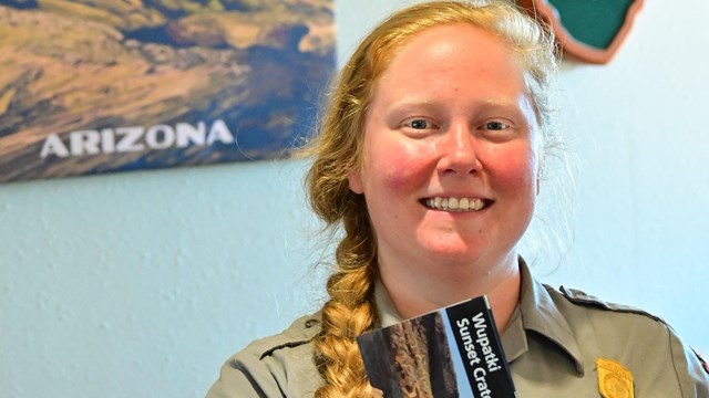 A smiling park ranger holds a brochure in front of the NPS arrowhead logo. 