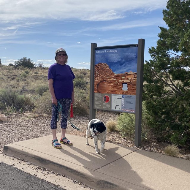 A smiling woman stands at a trail head with her dog on a leash. 