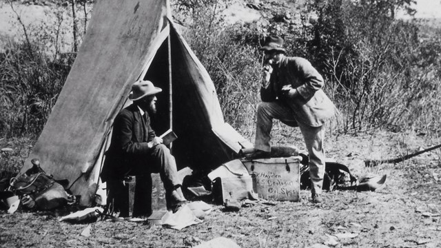 Man sits on a box in front of a canvas tent while another man stands next to him.