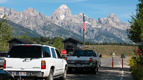 Two vehicles in line at a wooden kiosk with American flag next to it and mountains in the background