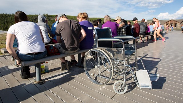 A wheelchair rests on the boardwalk behind a crowd of people sitting on benches.