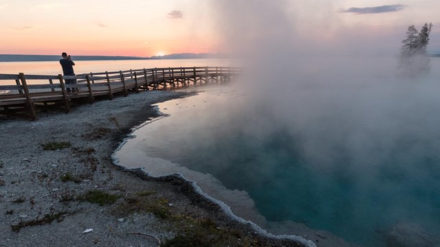 A person walks on a boardwalk near a hot spring at sunrise.