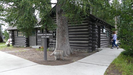 A visitor strolls across a concrete path toward the small wood-log building.