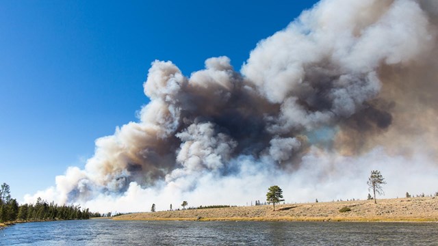 Large plume of wildfire smoke rising into the sky on the far side of a river