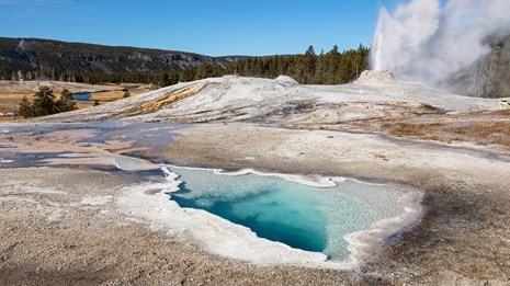 A blue hot spring with a geyser erupting in the background