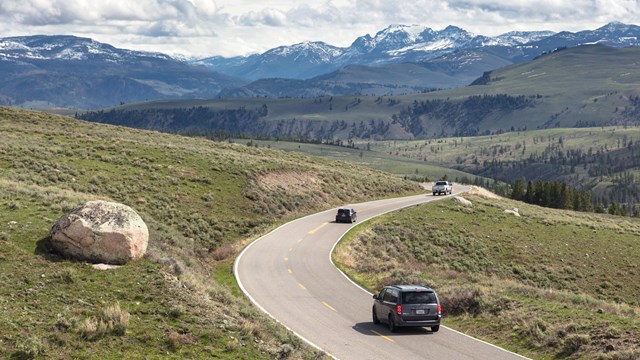Cars drive along a paved road through a meadow with snow-capped mountains in the background.
