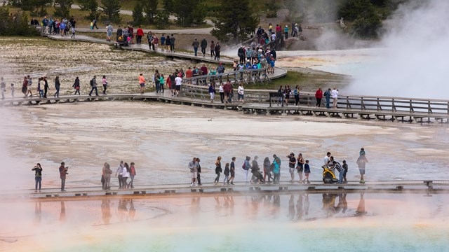 People walk across a boardwalk over steaming, colorful hot springs.
