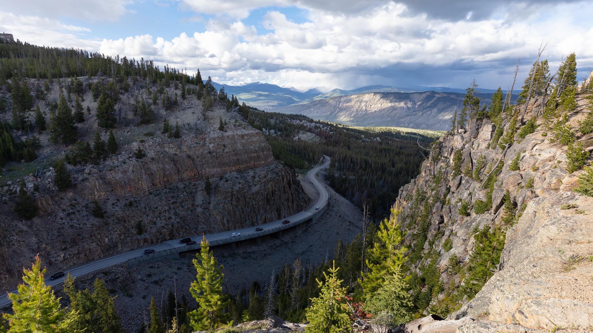Cars drive on a bridge that hugs the edge of a large cliff through the mountains.