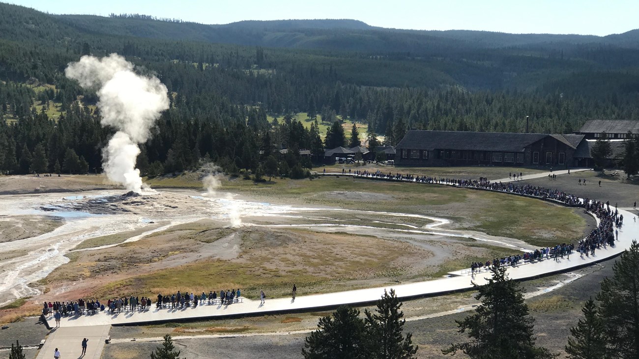 View from the top of a building shows visitors standing in a wide arc around a steaming geyser.