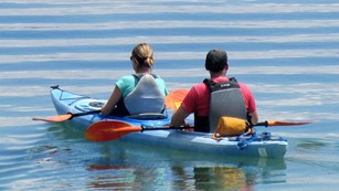 A couple paddles a two-person kayak across a still lake.
