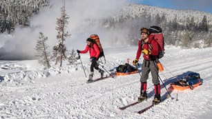 Two skiers traveling across a wide trail with backpacks to ski-sleds.