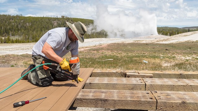 a park ranger nailing down new boards on a boardwalk in front of a geyser