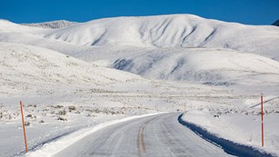 A thin layer of snow on a paved road with a snow-covered mountain in the background.