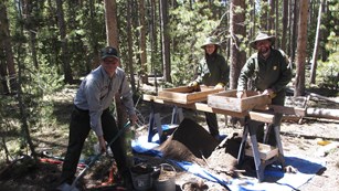 Three uniformed people working around excavated dirt. Two stand by wooden boxes; one uses a shovel.