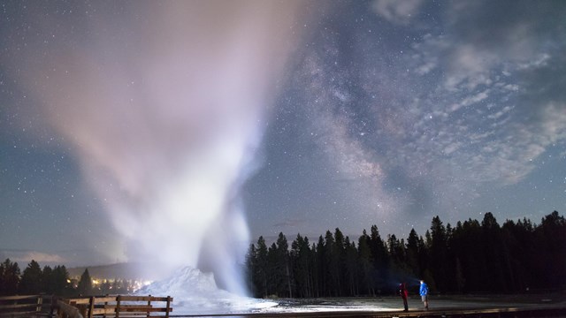 Two people watch a geyser erupt under a star-filled night sky.