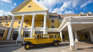 An old-fashioned yellow and black bus is parked in front of a tall, yellow inn with white columns.
