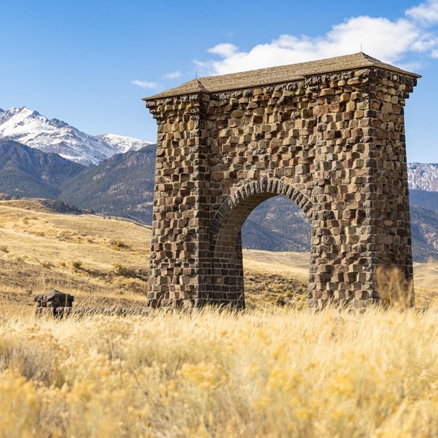 A large stone arch, Roosevelt Arch, stands in the foreground of a snow-capped mountain.