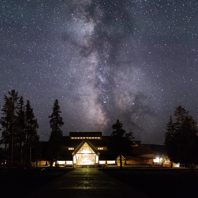 The milky way in the night sky rises above a lighted building, surrounded by tall trees.