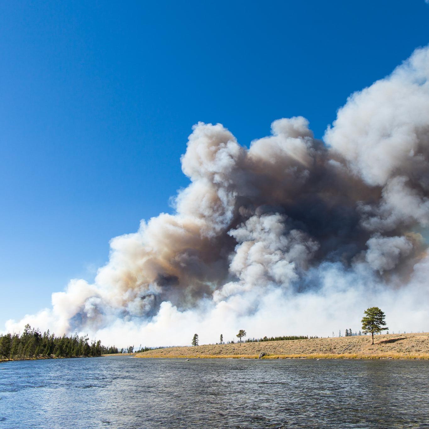 Large plume of wildfire smoke rising into the sky on the far side of a river