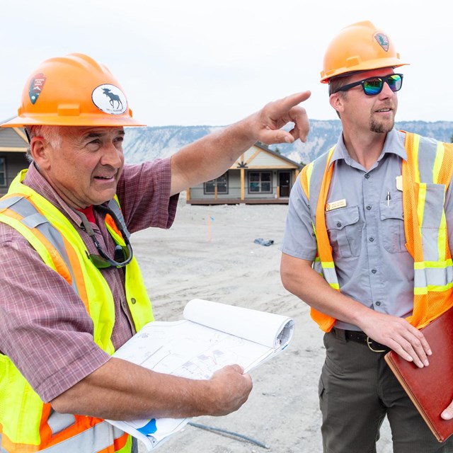 Two park service employees wearing construction vests and hard hats on a construction site