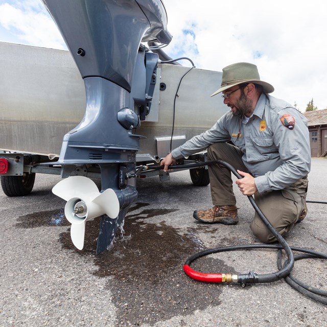 A park ranger with a dark beard and wearing a green and grey uniform kneels with a hose next a boat.