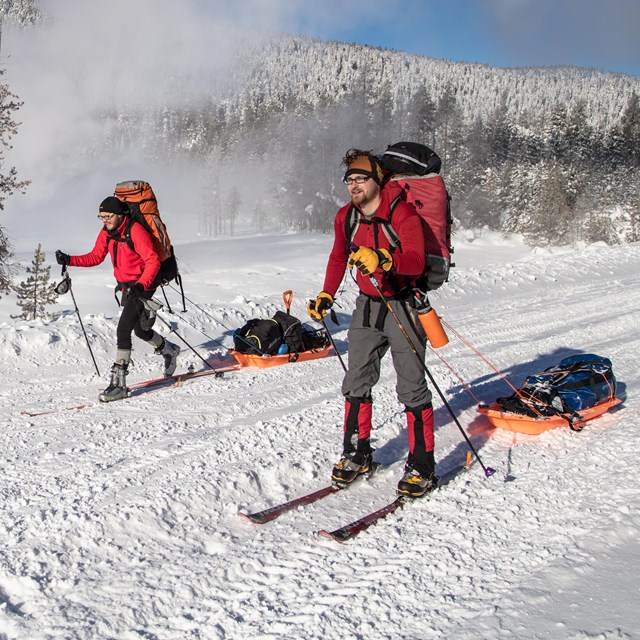 Two skiers traveling across a wide trail with backpacks to ski-sleds.