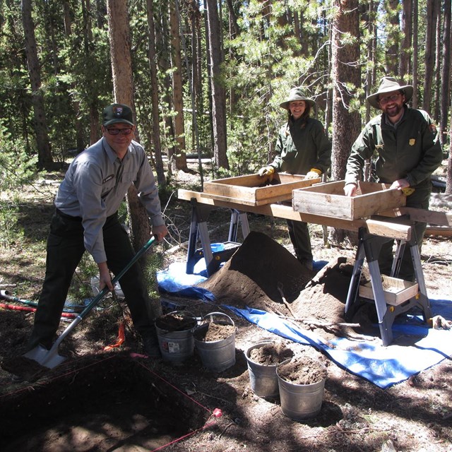 Three uniformed people working around excavated dirt. Two stand by wooden boxes; one uses a shovel.