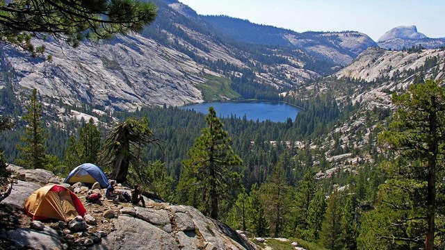 Two tents and campers overlook the high country, with Half Dome and a lake in the background.