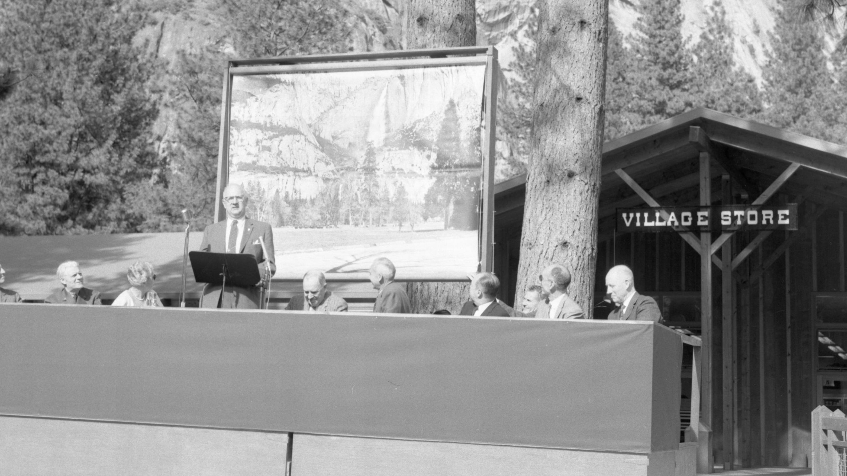 A man speaks on a temporary stage surrounded by other men; a scenic painting is behind him