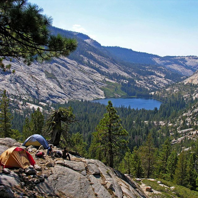 Two tents and campers overlook the high country, with Half Dome and a lake in the background.