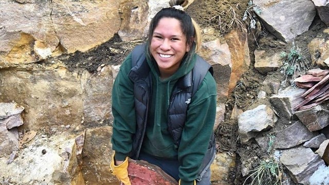 A young woman wearing a green jacket and black bookbag holds a red rock 