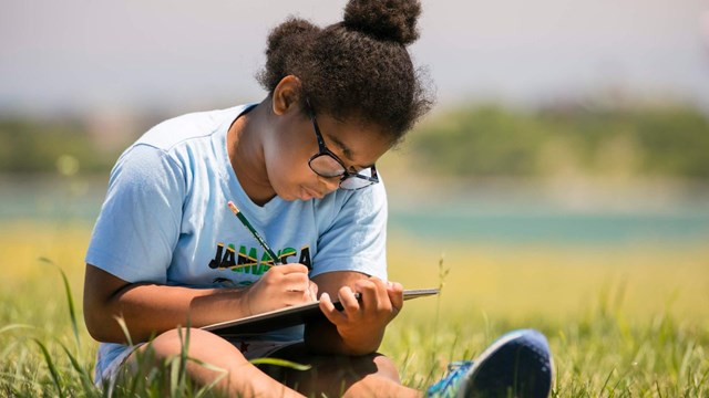 Girl sits in a field writing on her tablet