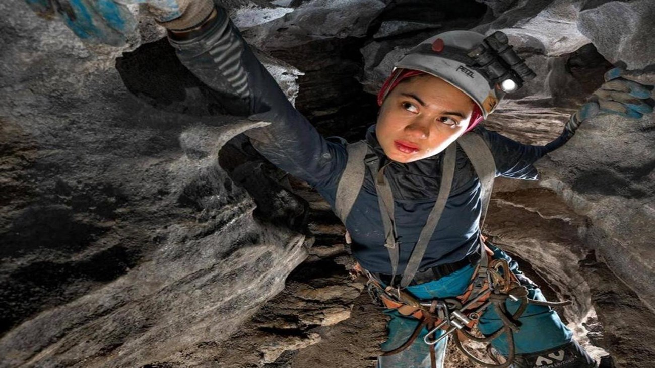 A young woman wearing a caving helmet and harness reaches forward to grab a handhold while caving.