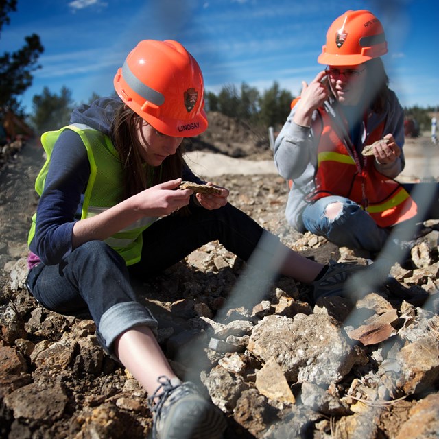 2 young women in safety vests and hard hats sit among a pile of rocks and look closely at rocks for 