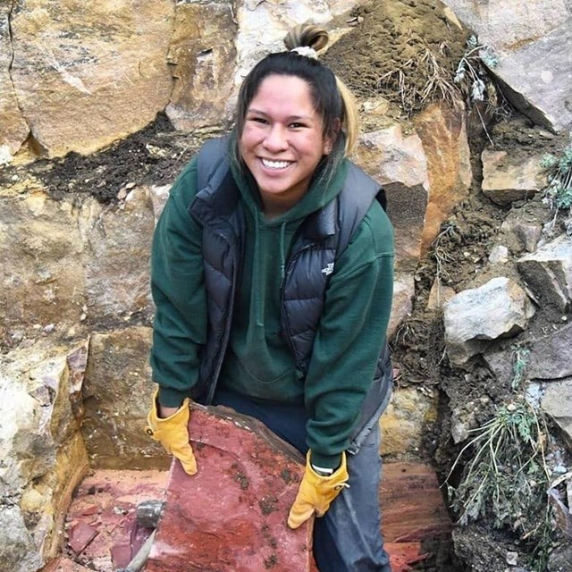 A young woman wearing a green jacket and black bookbag holds a red rock 